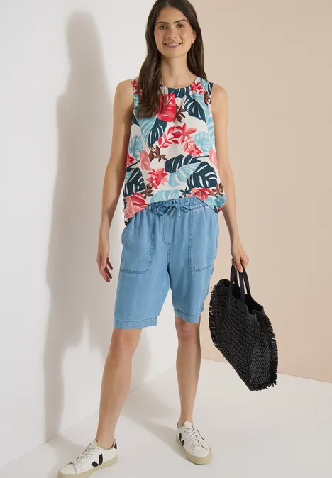 Blouse met ronde hals en bladerenprint light alabaster white Vrouw in mouwloos bloemen-top, losse blauwe shorts, witte sneakers en grote zwarte tas.