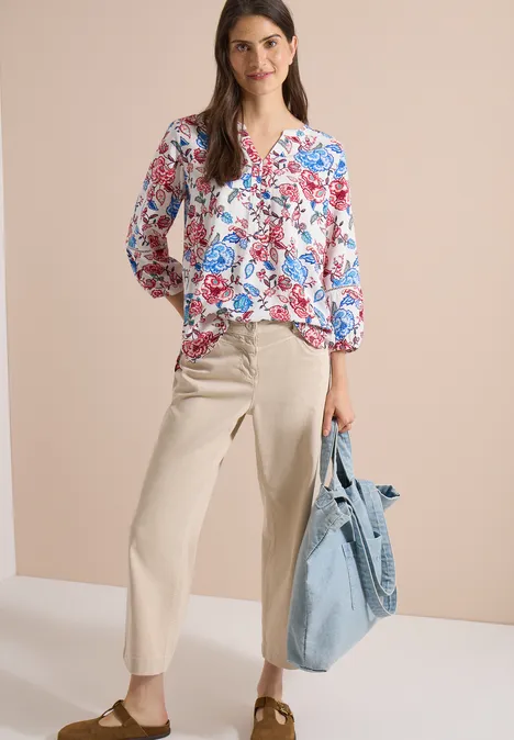 Vrouw met gebloemde blouse, beige wijde broek, bruine slippers en lichtblauwe tas voor lichte achtergrond.