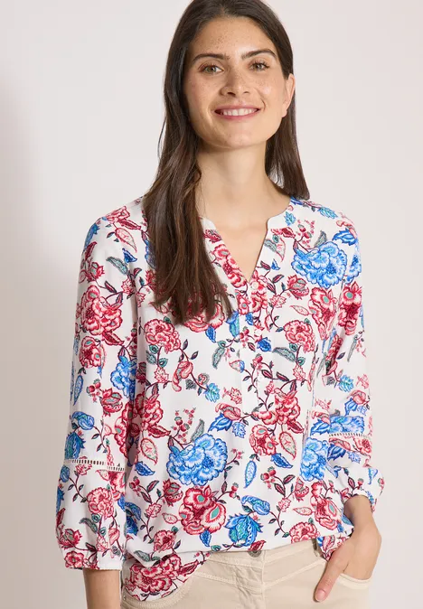 Vrouw draagt witte blouse met blauw-rood bloemenpatroon, V-hals en lange mouwen.