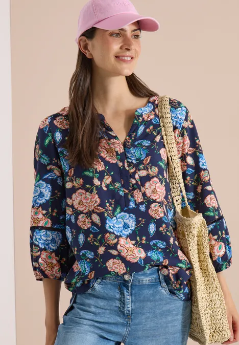 3/4-Arm Tunika mit floralem Muster urban dark blue Frau mit Blumenshirt, blauen Jeans, Strohtasche und rosa Cap vor hellem Hintergrund.