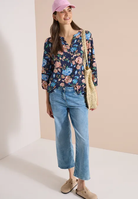 3/4-Arm Tunika mit floralem Muster urban dark blue Eine Frau trägt eine lockere, blau florale Bluse, gerade Jeans, Basttasche und beige Loafers vor hellem Hintergrund.