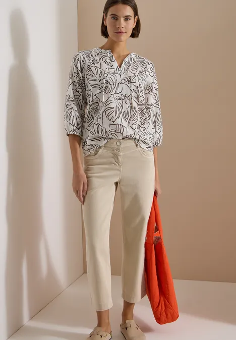 3/4-Arm Tunika mit Blätterprint light alabaster white Person trägt weiße, gemusterte Bluse, helle Stoffhose und hält eine orangefarbene Tasche vor beigem Hintergrund.