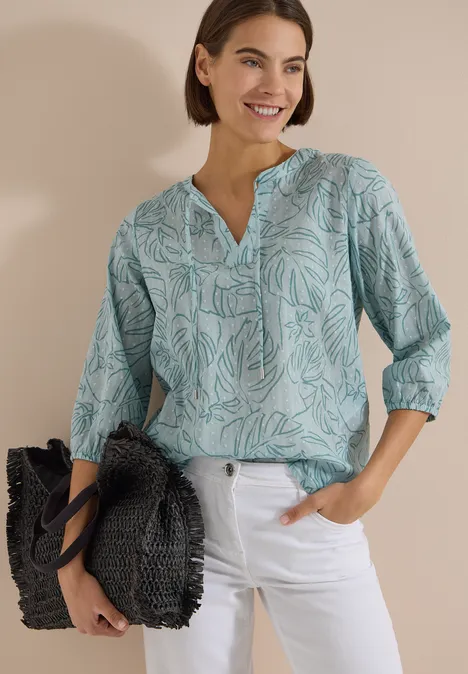 3/4-Arm Tunika mit Blätterprint crystal mint Frau trägt türkise, gemusterte Bluse mit 3/4-Ärmeln, weiße Jeans und schwarzen Flechtbeutel vor hellem Hintergrund.