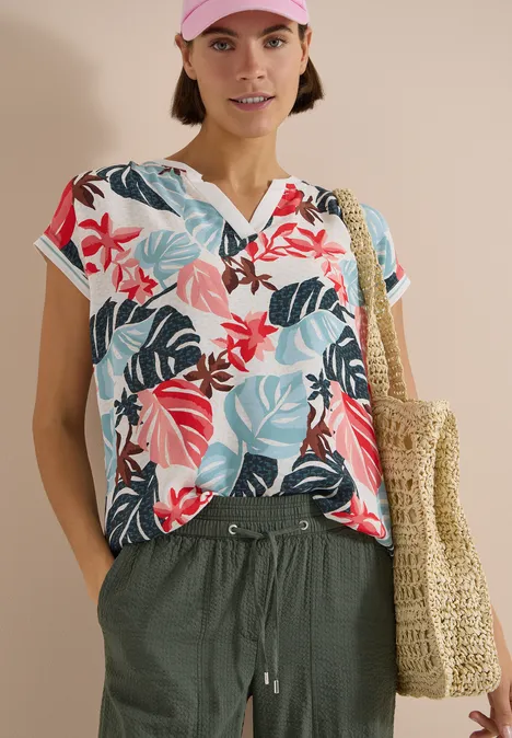 Bluse mit Split Neck und Printmix light alabaster white Eine Frau trägt ein buntes Blusen-Shirt mit tropischem Muster, grüne Hose und geflochtene Tasche.