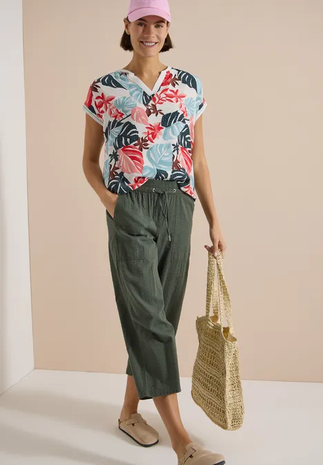 Bluse mit Split Neck und Printmix light alabaster white Frau trägt buntes, florales Top, weite grüne Hose und hält eine Strohtasche.