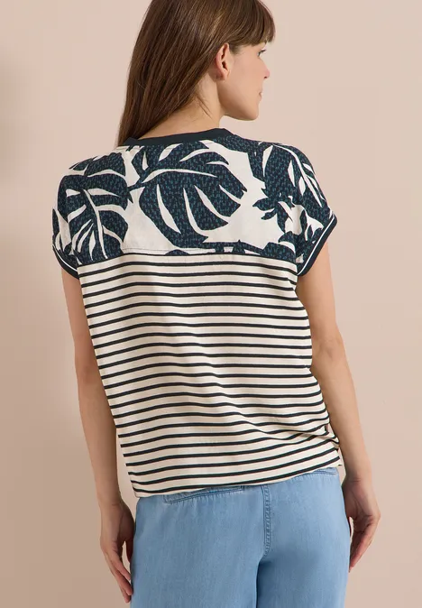 Blouse met gespleten hals en printmix palm breeze green Vrouw van achteren in een gestreept shirt met bladmotief en lichtblauwe broek voor een beige achtergrond.