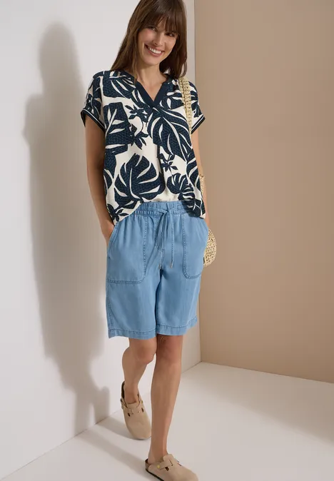 Blouse met gespleten hals en printmix palm breeze green Vrouw in blouse met bladmotief, losse jeansshorts, espadrilles en een ronde strooien tas.