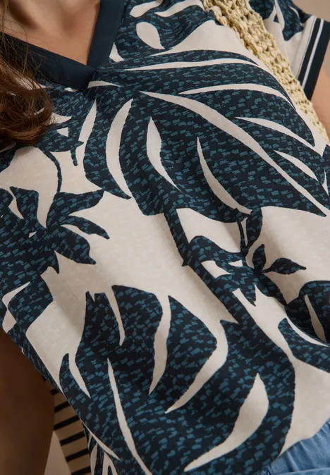 Blouse met gespleten hals en printmix palm breeze green Close-up van een blouse met donkerblauwe monstera bladerenprint op een beige achtergrond.