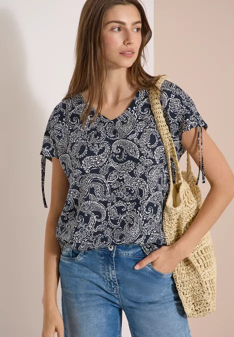 Femme en chemisier à motif cachemire bleu, jean et sac en paille tressée.