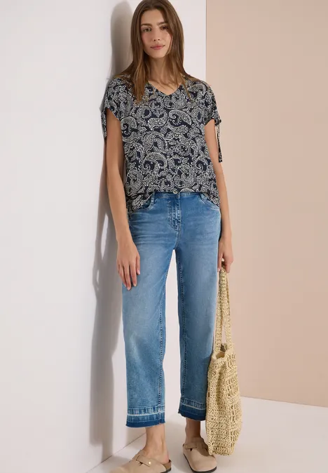 Femme portant un jean droit, un haut à manches courtes à motif et un sac tressé, devant un mur clair.