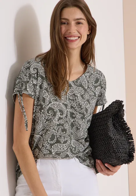 Overhemd met gerimpeld detail khaki Frau in grüner Paisley-Bluse mit Kurzärmeln, weißen Hosen und schwarzer gewebter Clutch.