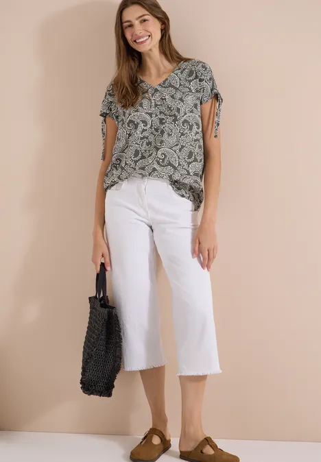 Overhemd met gerimpeld detail khaki Frau trägt gemustertes Top, weiße Culotte, braune Slipper und hält eine schwarze Tote-Bag, neutraler Hintergrund.