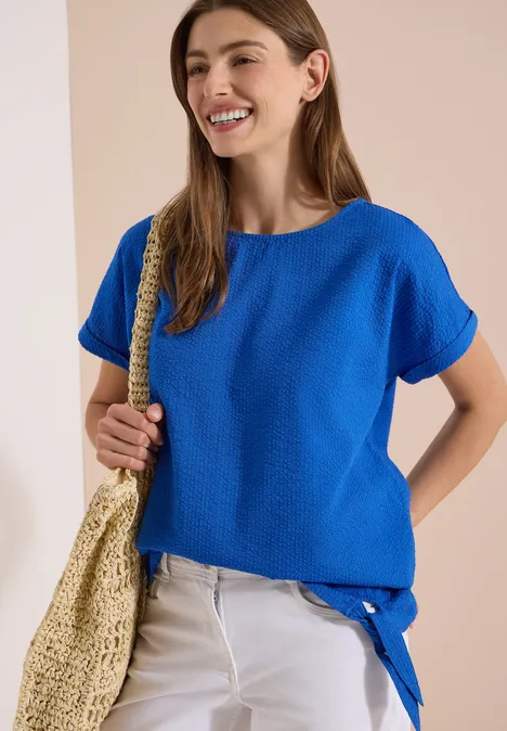Blouse met ronde hals en knoopdetail surf blue Frau in lockerem, kobaltblauem Oberteil mit Knotendetail, weißen Jeans und Strohtasche vor neutralem Hintergrund.