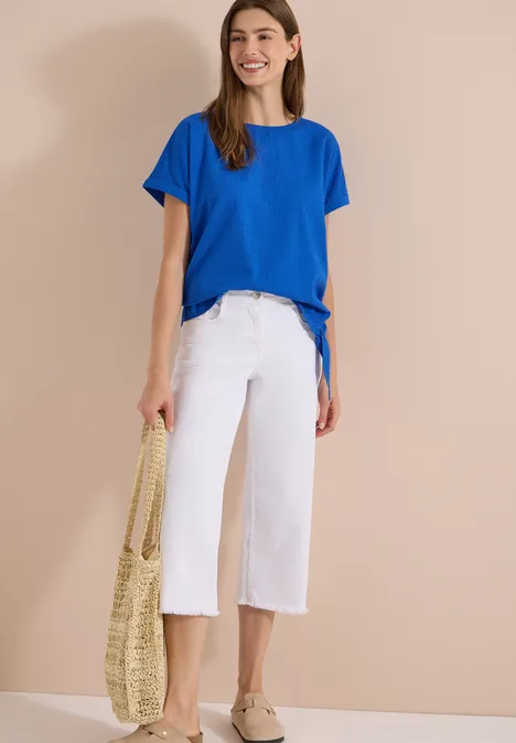 Blouse met ronde hals en knoopdetail surf blue Frau steht vor beigem Hintergrund, trägt blaues T-Shirt, weiße Culottes, Espadrilles und eine geflochtene Tasche.
