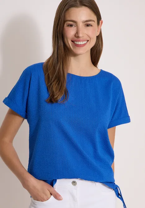 Blouse met ronde hals en knoopdetail surf blue Frau in blauem Kurzarmshirt mit Rundhalsausschnitt steht vor hellem Hintergrund.
