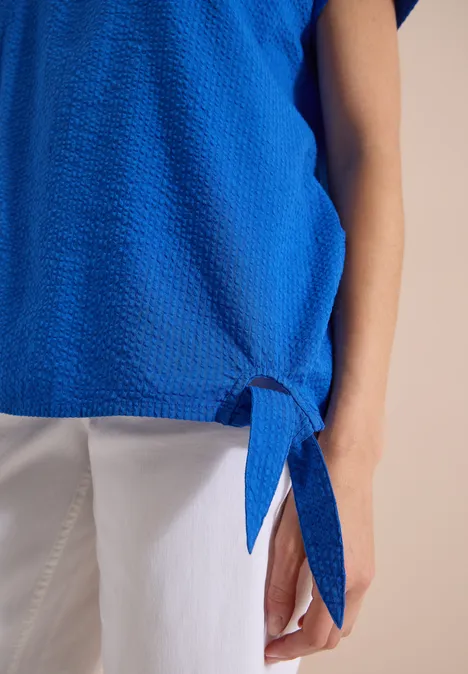 Blouse met ronde hals en knoopdetail surf blue Nahaufnahme einer Person in blauem Oberteil mit Knoten am Saum und weißer Hose.