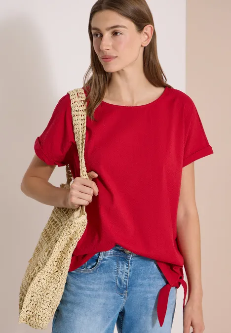 Frau in rotem T-Shirt mit seitlicher Knoten, blauer Jeans und Basttasche über der Schulter.