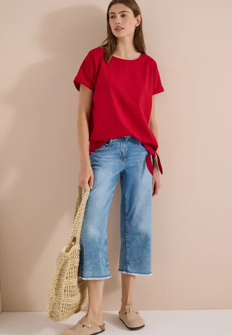 Frau in rotem T-Shirt, blauen Culotte-Jeans, beigen Slip-ons hält eine geflochtene Tasche vor beigem Hintergrund.