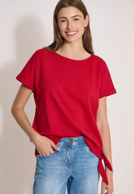Frau trägt rotes T-Shirt und Jeans, steht vor weißem Hintergrund.