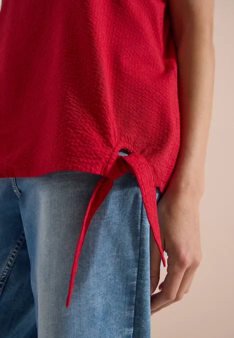 Rote, texturierte ärmellose Bluse mit abgenähtem Saum und Kordelzug, kombiniert mit blauen Jeans.