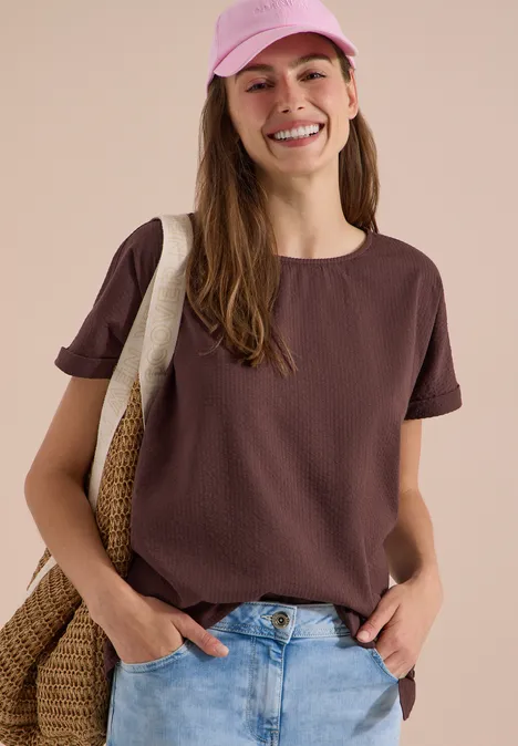 Blouse met ronde hals en knoopdetail brown cacao Eine junge Frau trägt ein braunes T-Shirt, Jeans und eine rosa Cap, eine Strohtasche über der Schulter.