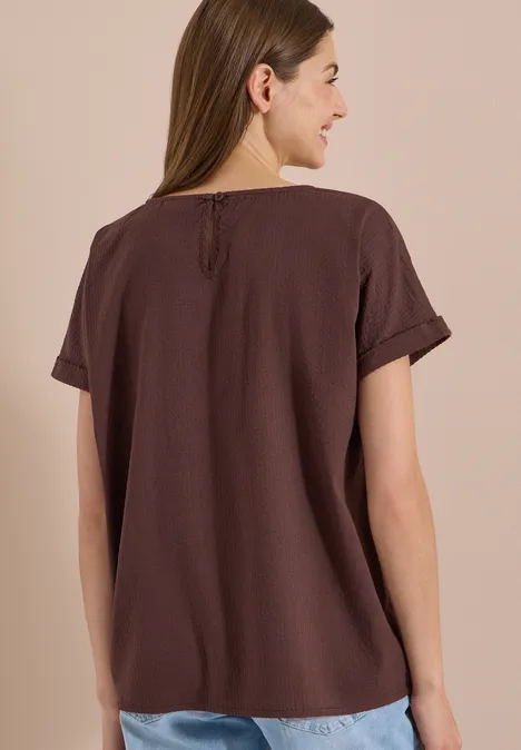 Blouse met ronde hals en knoopdetail brown cacao Rückenansicht einer Frau in einem schlichten braunen T-Shirt mit umgeschlagenen Ärmeln.