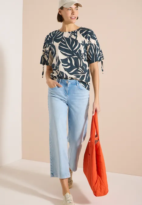 Bluse mit Rundhals und Raffung palm breeze green Frau trägt eine Bluse mit Blättermuster, helle Schlagjeans, grüne Sneaker und rote Tasche.
