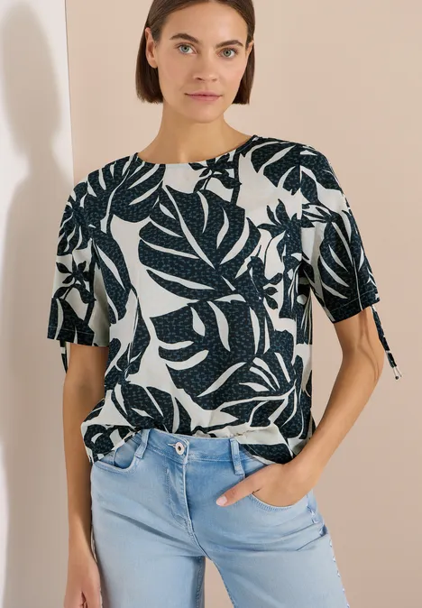 Bluse mit Rundhals und Raffung palm breeze green Frau in weißem Kurzarmshirt mit dunkelgrünem Blätterprint, locker in hellblaue Jeans gesteckt, vor beigem Hintergrund.
