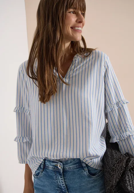 Frau trägt locker sitzende, blau-weiß gestreifte Bluse und Jeans, hält grauen Strickpullover.