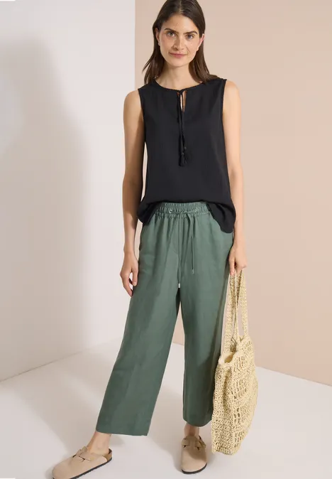 Blusentop mit Split Neck und Bändern Black Frau trägt schwarzen ärmellosen Top, weite grüne Hose, beige Schuhe und eine Netz-Tasche.