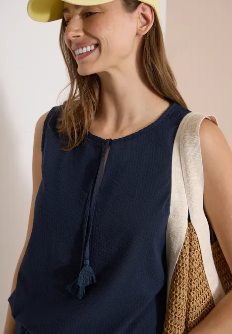 Femme en top bleu marine sans manches à pompons, portant deux sacs d’épaule en toile et paille.