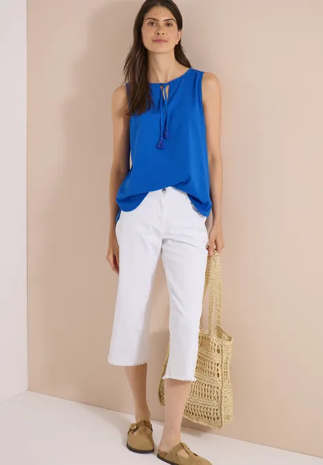 Blusentop mit Split Neck und Bändern surf blue Frau mit blauem Top, weißen Culotte-Hosen und Strohtasche, trägt Sandalen.