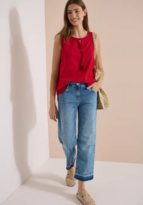 Femme en top rouge sans manches, jean bleu ample et sac tressé en paille devant un mur clair.