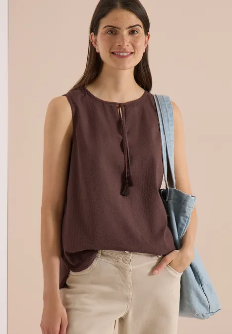 Blusentop mit Split Neck und Bändern brown cacao Frau trägt ärmelloses, braunes Top mit Quasten, helle Hose und blaue Jeans-Tasche über der Schulter.