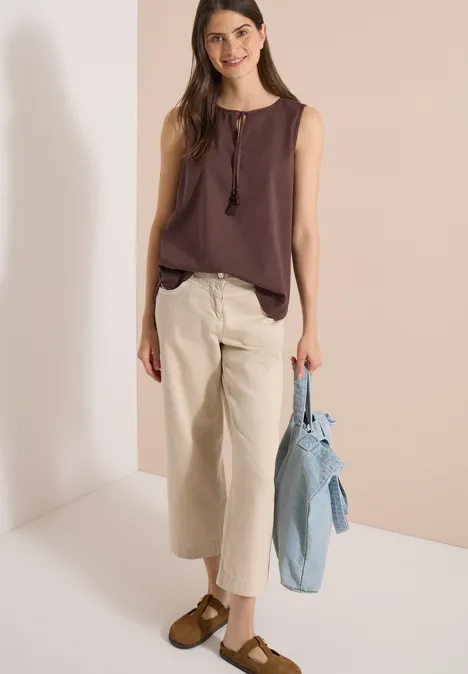 Blusentop mit Split Neck und Bändern brown cacao Frau in braunem Top, heller Culotte-Hose und braunen Schuhen, hält hellblaue Jeansjacke.