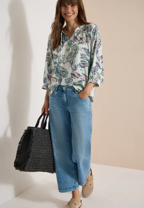 Vrouw met bloemprint blouse, wijde blauwe jeans en zwarte gevlochten tas, casual en stijlvol.
