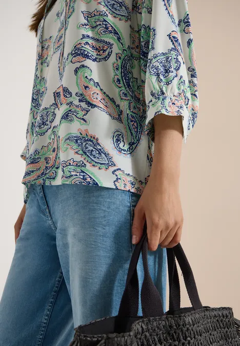 Persoon in blouse met paisleypatroon en spijkerbroek houdt een zwarte gevlochten tas vast.