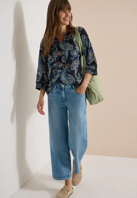 Vrouw in gebloemde blouse, wijd jeans, enkellaarsjes en groene tas voor een lichte muur.