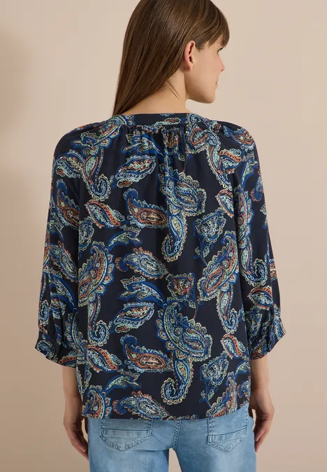 Vrouw van achteren in een donkerblauw blousje met paisley-print en spijkerbroek voor een beige achtergrond.