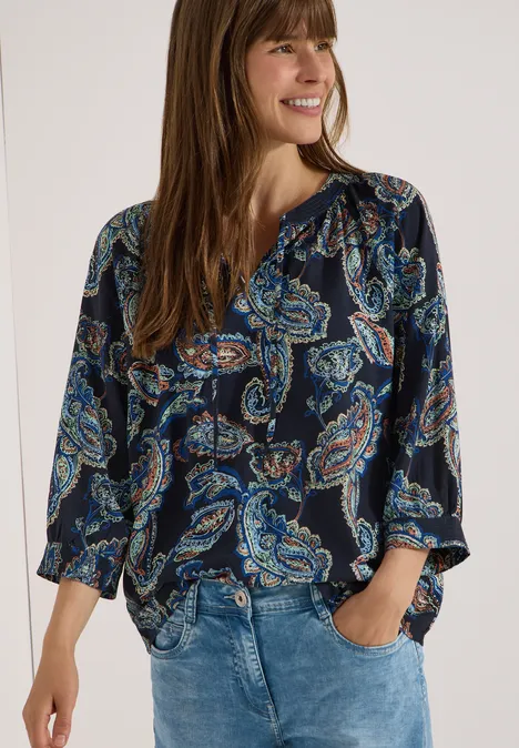 Vrouw in blauw blouse met paisley-patroon en jeans, handen in de zakken.