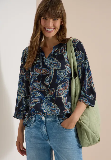 Vrouw in donker paisleybloes, jeans en groene tas, ontspannen pose.