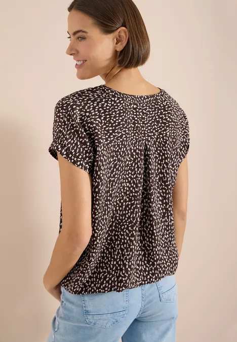 Blouse met hangende schouder en print brown cacao Rugaanzicht van een vrouw in een zwart, gestippeld shirt met korte mouwen en lichtblauwe jeans.