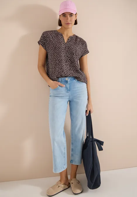 Blouse met hangende schouder en print brown cacao Vrouw draagt een kort mouw blouse met patroon, lichtblauwe rechte jeans, beige loafers en een donkerblauwe shopper.