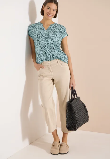 Frau lehnt in legerem Outfit mit gemusterter Bluse, beigen Hosen und schwarzer Tasche an Wand.
