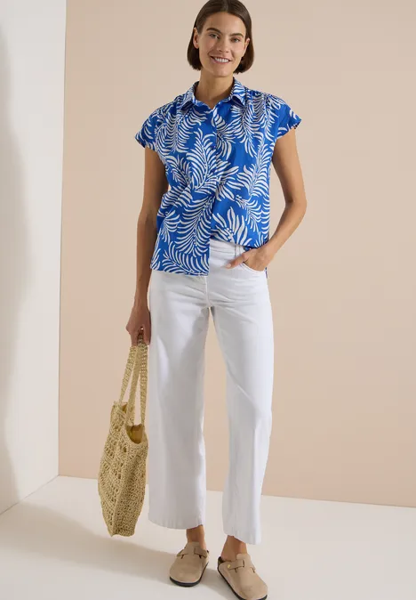 Kurzarm Hemdbluse mit Print surf blue Frau in blauem Blattmuster-Top, weißer Hose, Basttasche und flachen Schuhen vor neutralem Hintergrund.