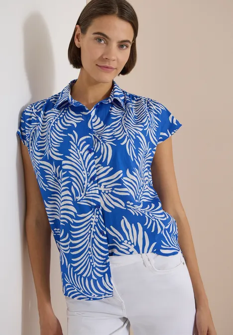 Kurzarm Hemdbluse mit Print surf blue Frau in tiefblauem Hemd mit weißem Blattprint und weißer Hose lehnt an schlichtem hellen Hintergrund.