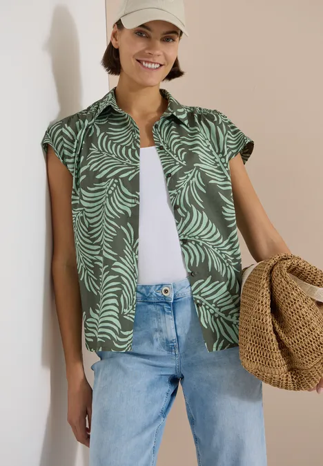 Kurzarm Hemdbluse mit Print khaki Frau trägt grünes, gemustertes Kurzarmhemd über weißem Top, hellblaue Jeans und hält Strohtasche.
