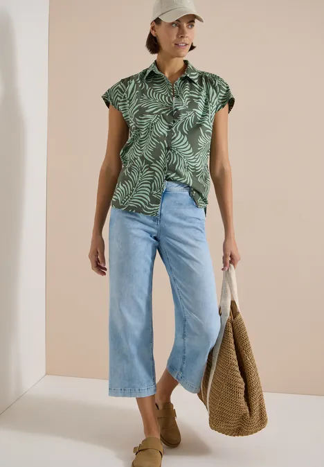 Kurzarm Hemdbluse mit Print khaki Frau mit grüner, gemusterter Kurzarmbluse, Cropped-Jeans, beigen Mules und Strohtasche vor hellem Hintergrund.
