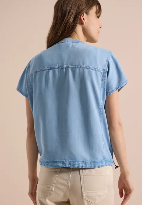 Frau von hinten in hellblauer, kurzärmeliger Jeansbluse und hellen Hosen.