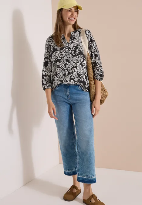 Femme en chemisier à motifs, jean bleu, sandales marron, sac en paille et chapeau jaune sur fond clair.